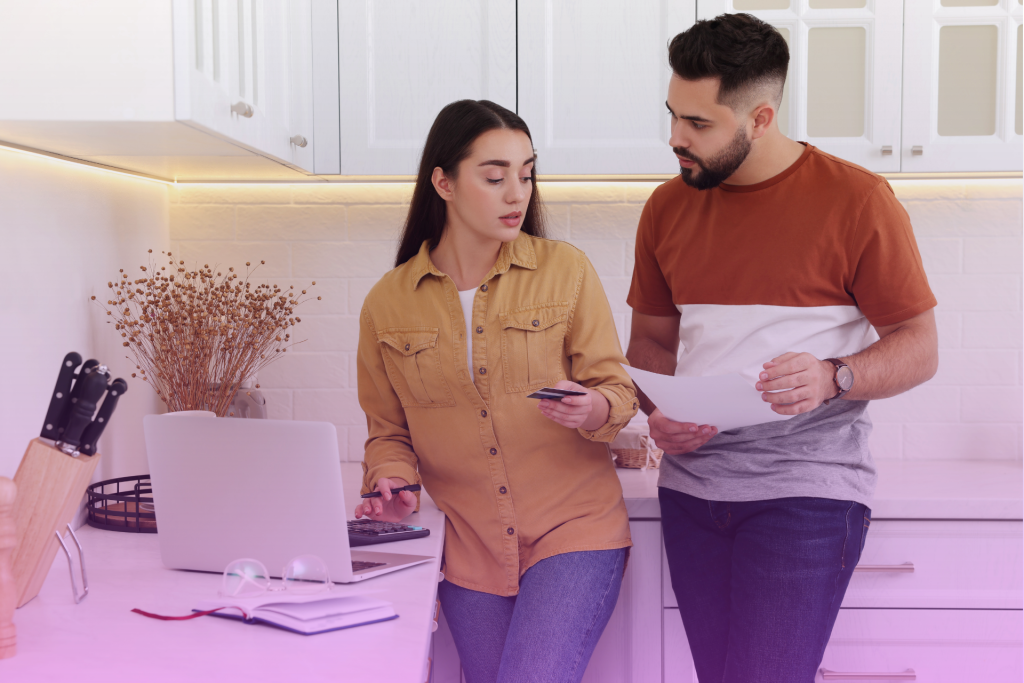 A couple sitting at a kitchen table, talking and looking at financial documents spread out in front of them.