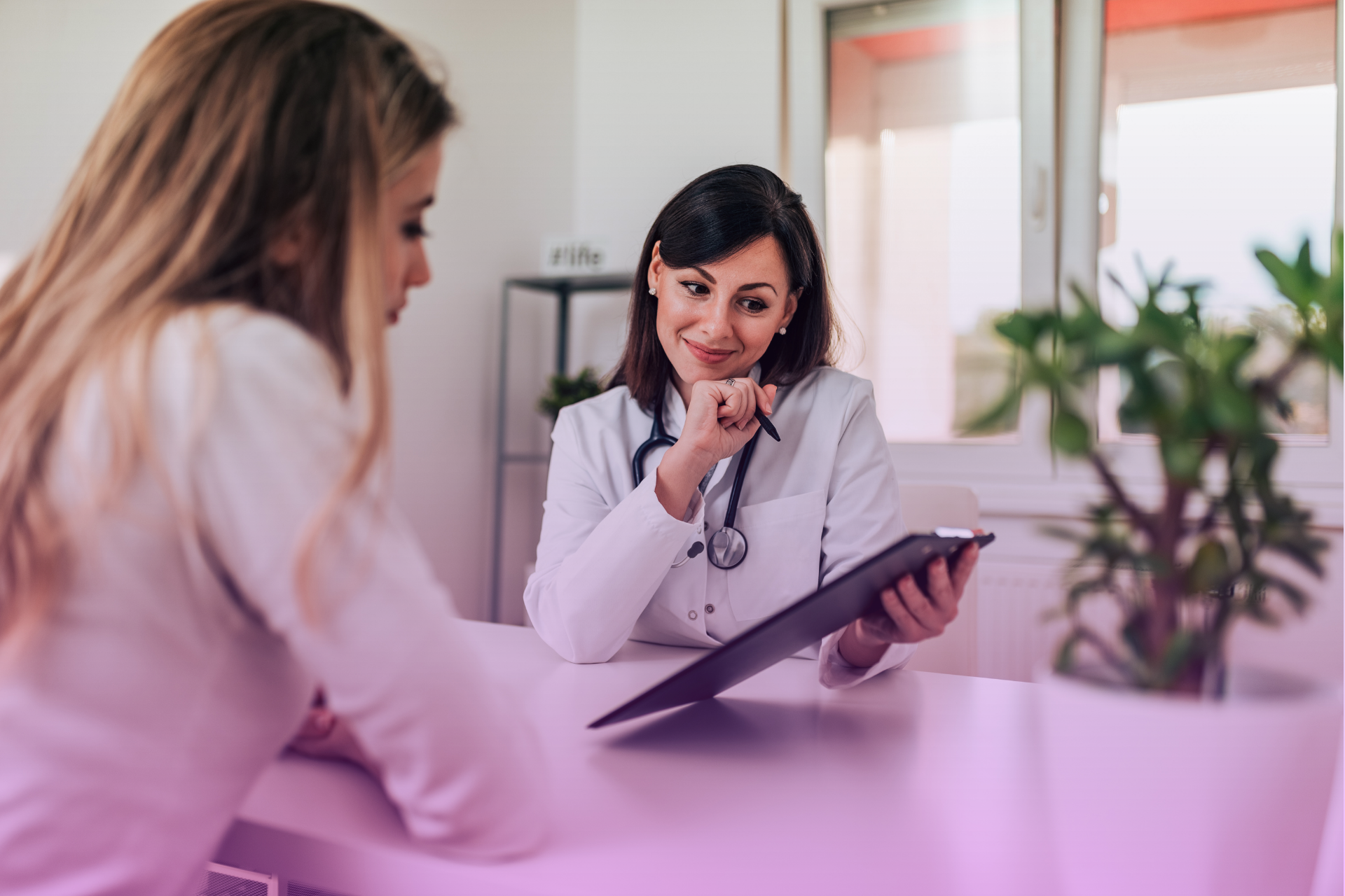 A kind-looking doctor in scrubs sits with a smiling patient, taking notes on a clipboard.