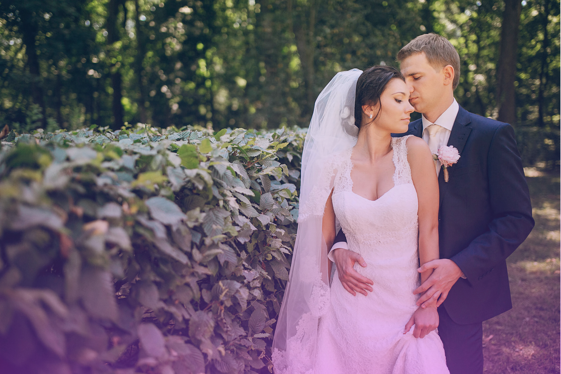 A couple posing together on their wedding day, dressed in formal attire, smiling and holding each other closely against a beautiful outdoor backdrop.