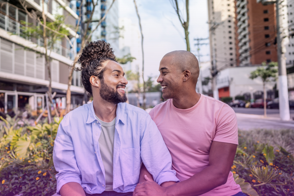 A gay couple sits on a park bench, smiling and engaged in a thoughtful conversation about prenuptial agreements, surrounded by lush greenery.