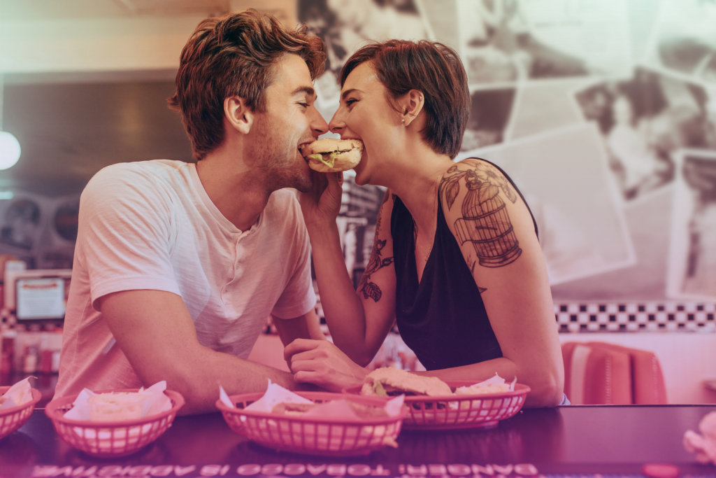 A couple laughing and gazing into each other's eyes while sharing a burger, capturing a moment of joy and connection.