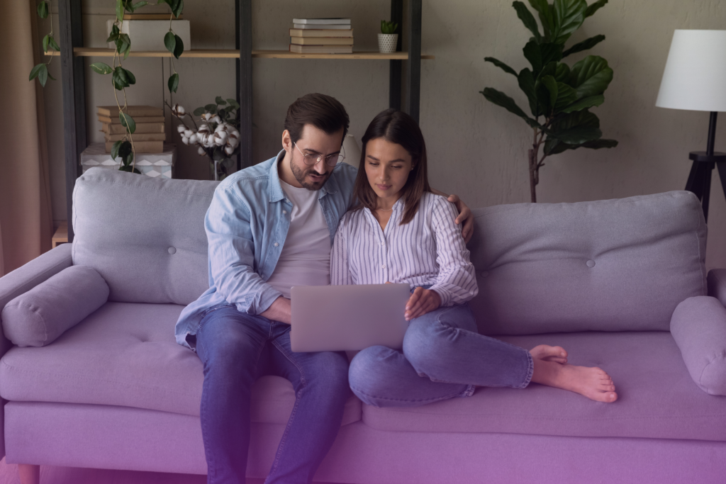 A couple sitting at a table with a laptop open, looking at each other with concerned expressions, possibly facing a disagreement about their postnup.