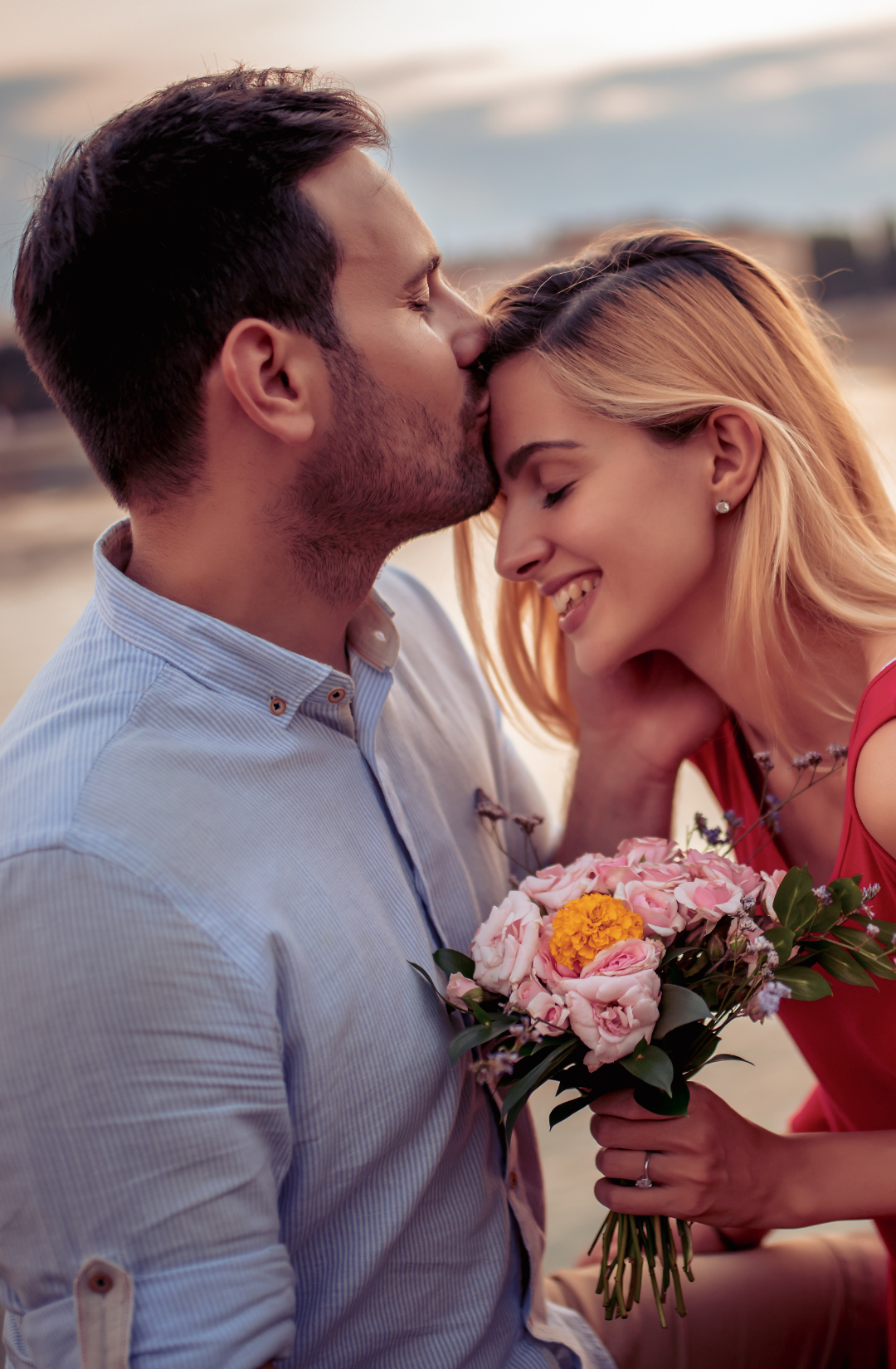 A romantic couple at sunset, where a man gently kisses the woman’s forehead. She smiles while holding a bouquet of flowers in her hand.