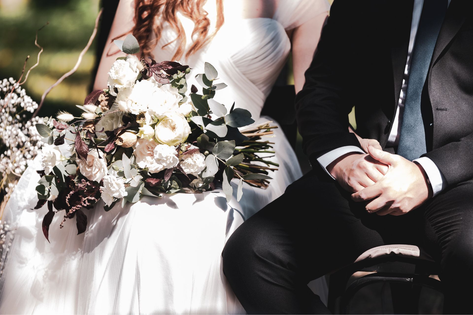 Close-up of a bride and groom sitting side by side, showing only their wedding attire and a floral bouquet, creating an intimate and elegant wedding moment.