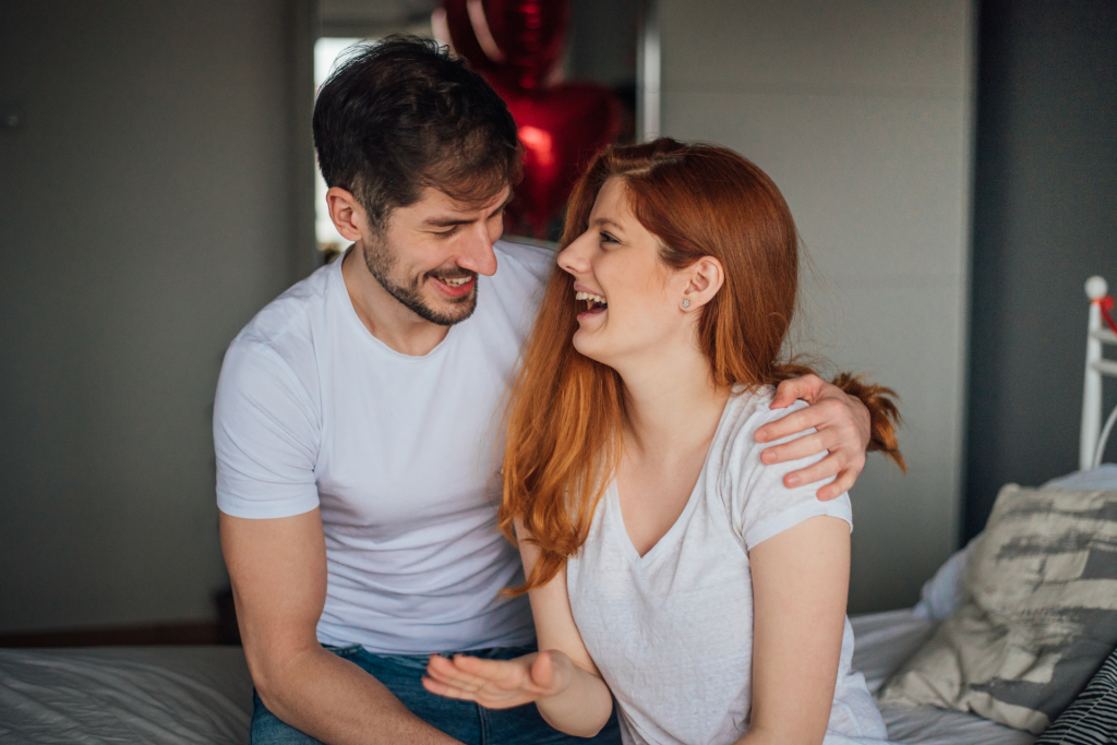 A couple sitting on the bed, laughing and looking into each other’s eyes, symbolizing a warm and open conversation.