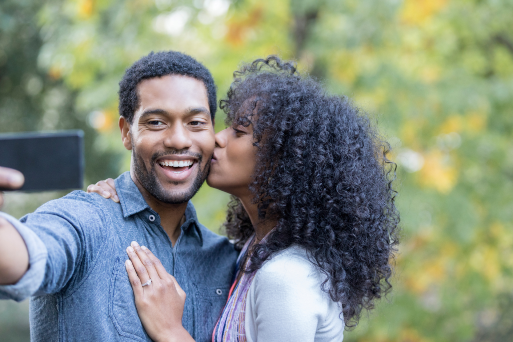 A couple taking a selfie while the woman kisses the man on the cheek and he holds the phone to capture the photo.