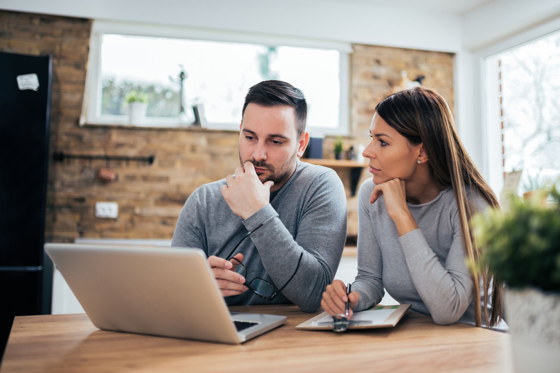 A worried couple reviews their finances at a table; the man types on a laptop while the woman looks on with concern, holding a pen and a notebook.