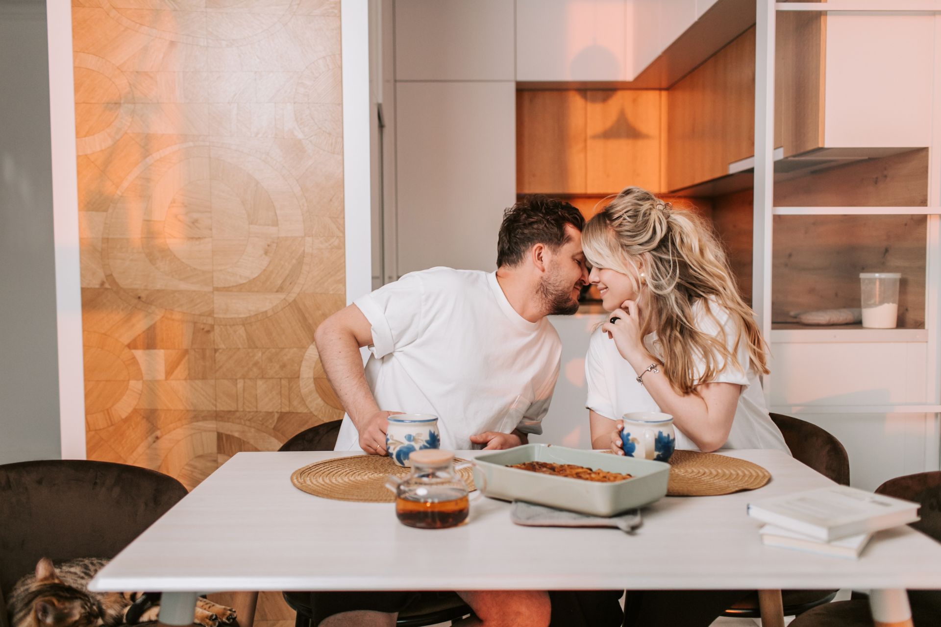 A couple holding coffee mugs, smiling and leaning in for a kiss while having breakfast together