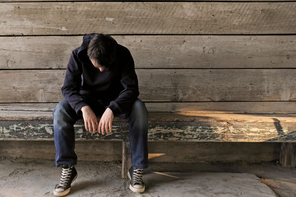 A solitary man sitting with his head lowered, gazing at the floor. His face is not visible, but his posture conveys deep sadness and loneliness.