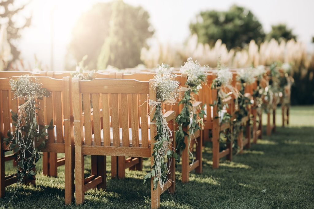 Rows of chairs decorated with floral arrangements at an outdoor wedding ceremony, set up in a natural open-air setting.