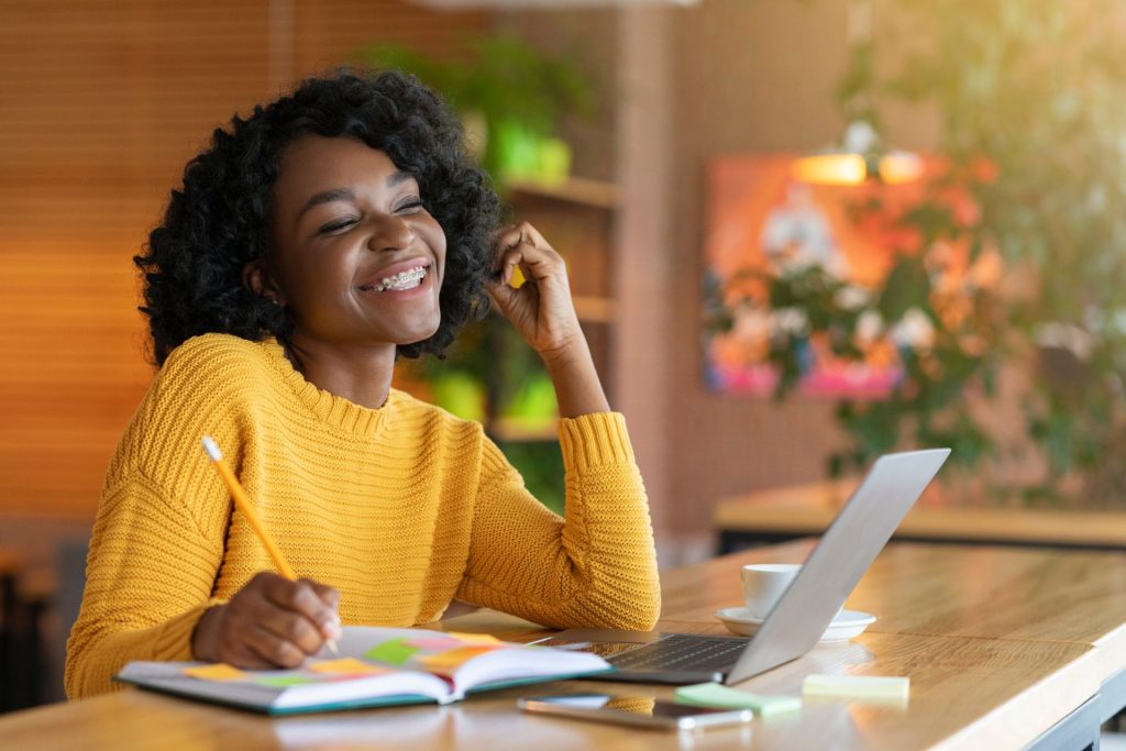 A smiling woman sits at her desk, talking on the phone while holding a pen and writing in a notebook filled with sticky notes, with her laptop open in front of her.