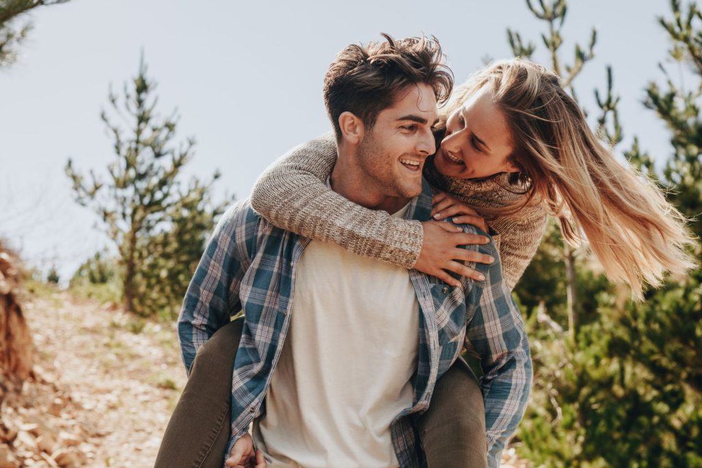 Energetic couple smiling widely as they playfully leap or jump together on a sunny day in a grassy park setting.