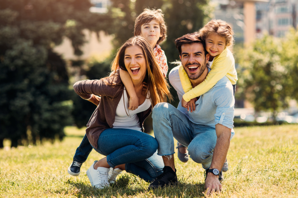 A happy family (mother, father, and children) laughing loudly while playing together in the bright sunshine at a park.