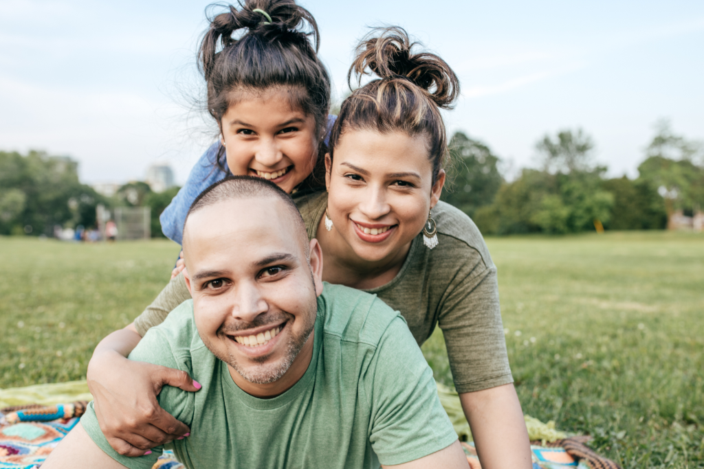 A happy mother, father, and daughter smiling and playing together on the grass in a sunny park.