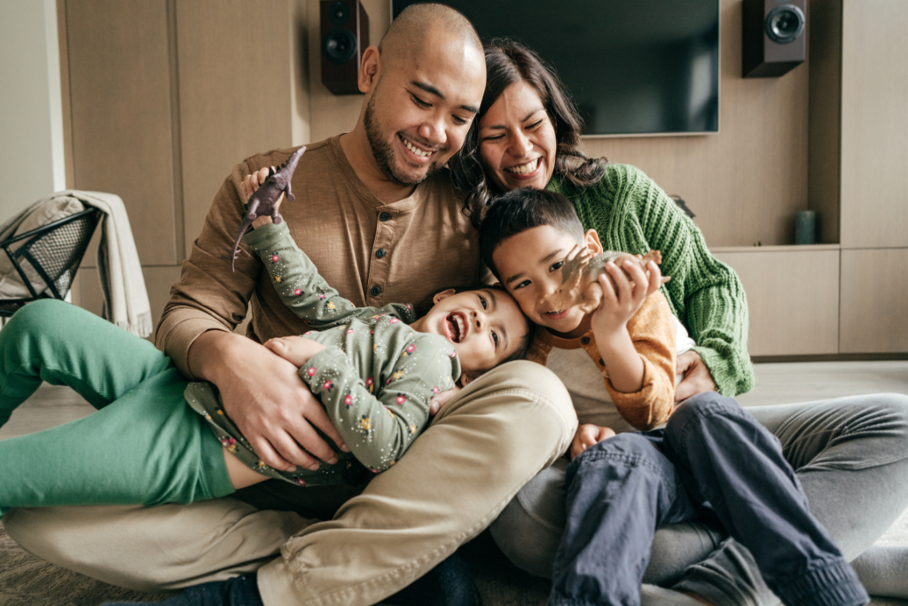 Joyful parents, son, and daughter laughing while playing together with toys on the floor inside their home.