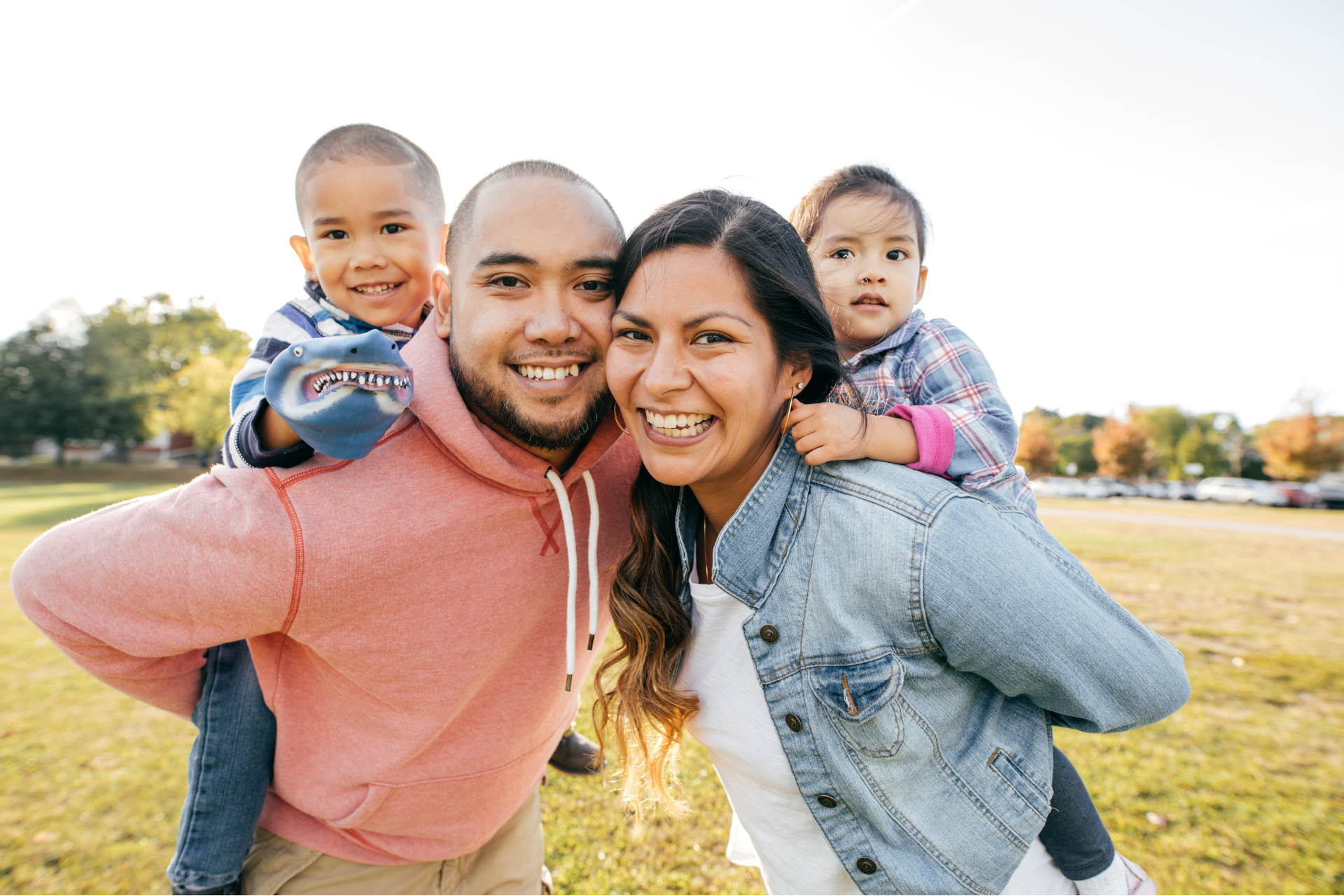 A smiling Latino family of four (mother, father, daughter, and son) posing directly at the camera in a sunny park setting.