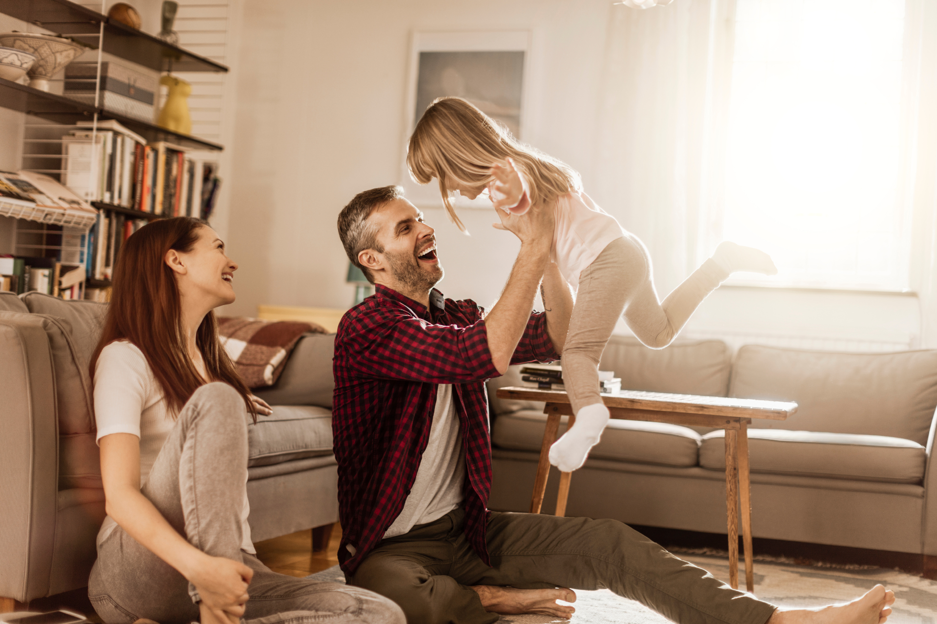 parents-daughter-playtime-laughter Happy family playing together indoors, with the father raising the daughter overhead, and the mother watching with a smile.
