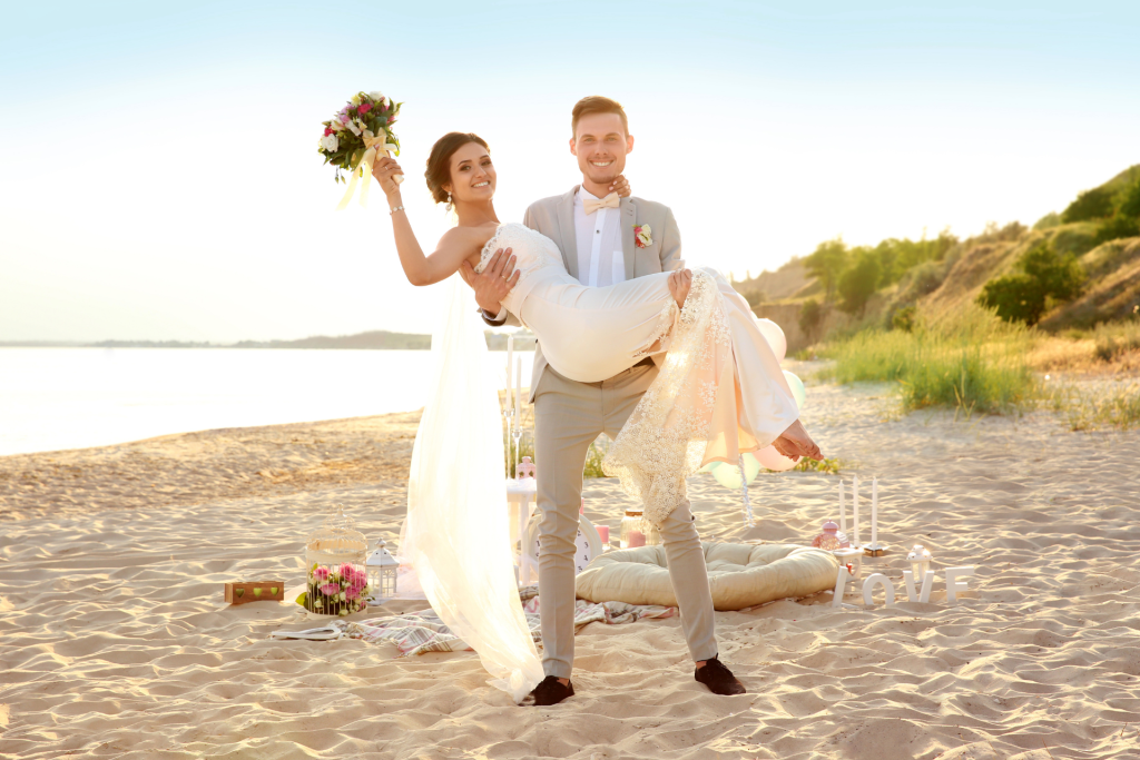 A couple celebrating their wedding on the beach, with the groom lifting the bride as she holds her bouquet, wedding decorations visible in the background, both smiling at the camera