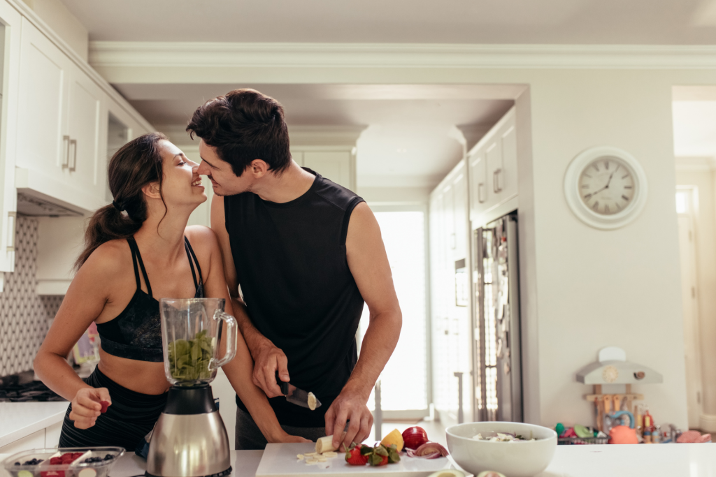 A couple cooking together in the kitchen, making a fruit smoothie while laughing and sharing a kiss