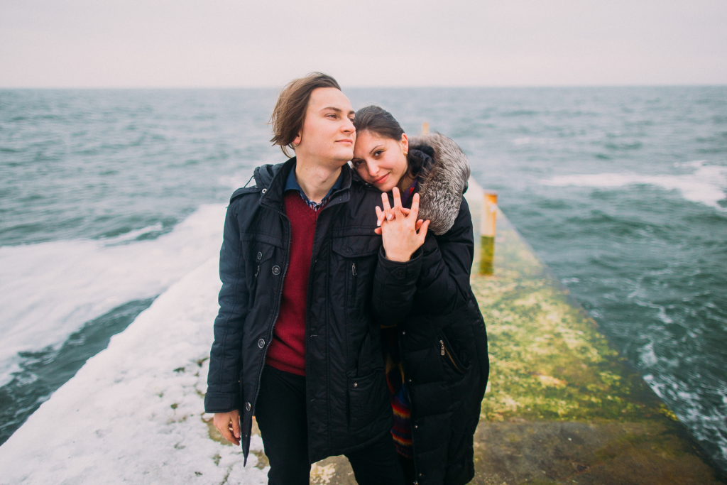 A couple standing at a harbor with the ocean in the background, holding hands and embracing while looking at the camera on a cold day