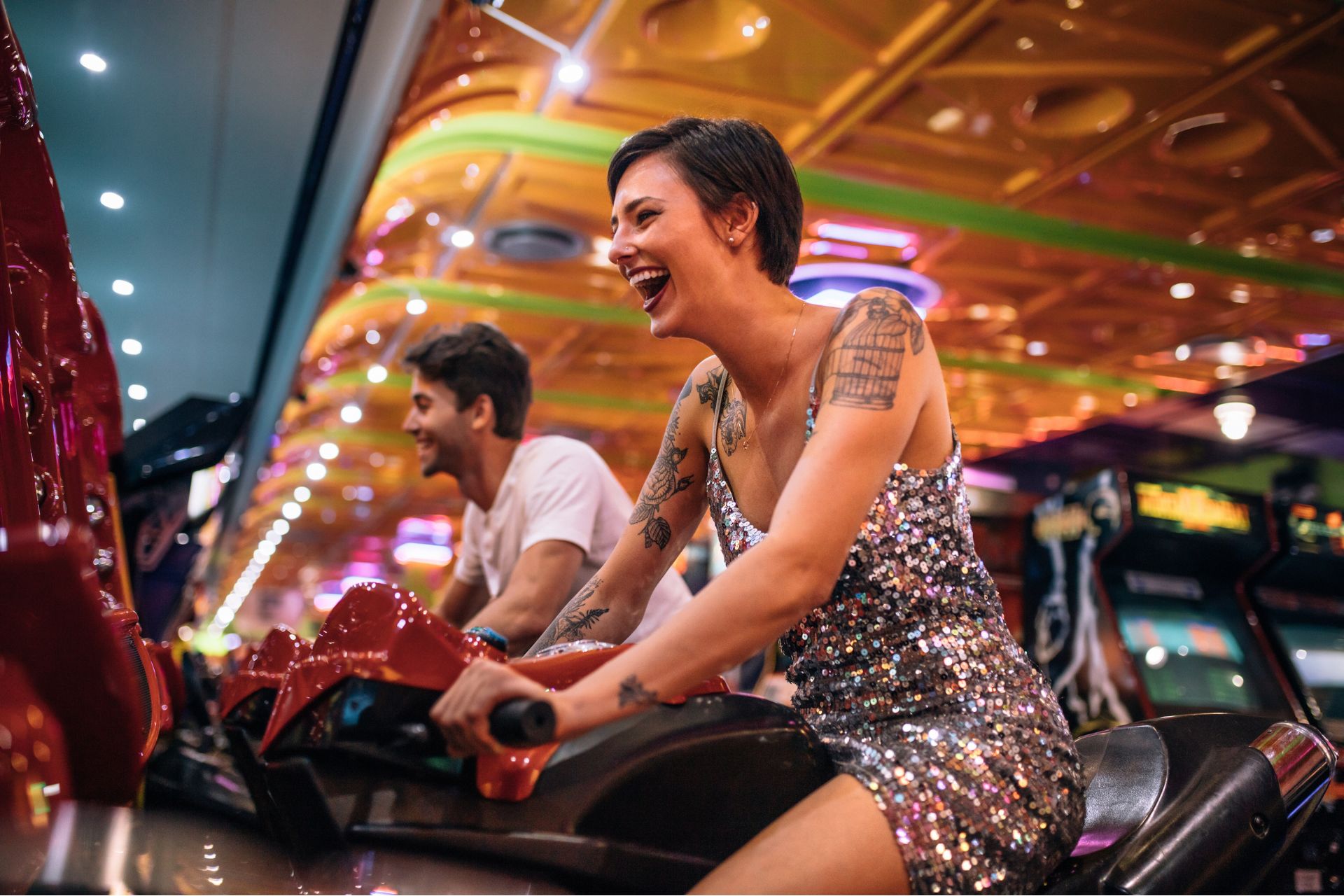 A couple laughing while playing a motorcycle racing arcade game, each riding a motion-based motorcycle game inside a casino