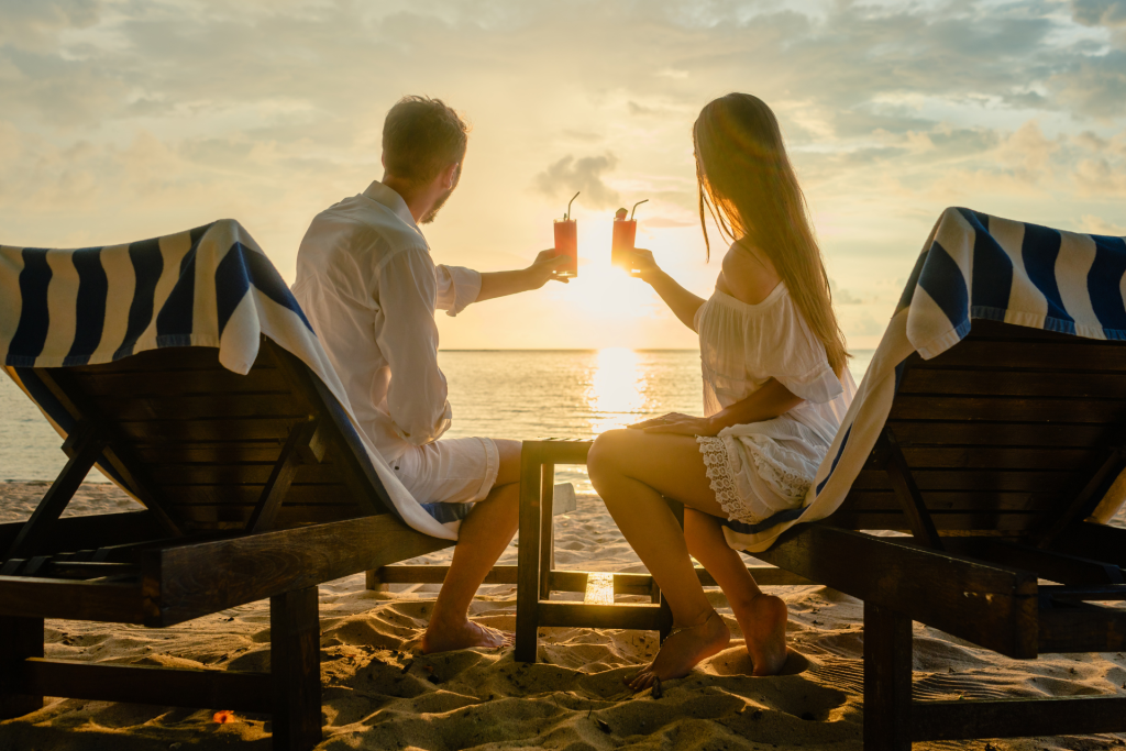 A couple sitting on the sand at the beach during sunset, seen from behind, toasting with cocktails while watching the sunset