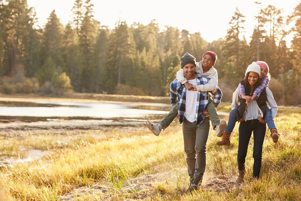 A family playing outdoors on a sunny but cold day, with each parent carrying a child on their back while everyone laughs and plays