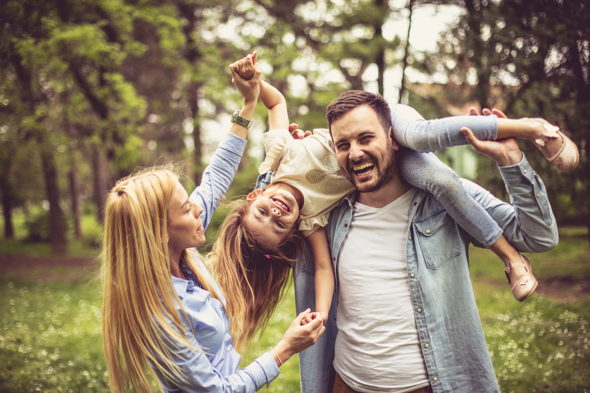 A family playing in a park on a sunny day, with the father lifting his daughter while the mother holds her hands and everyone is laughing