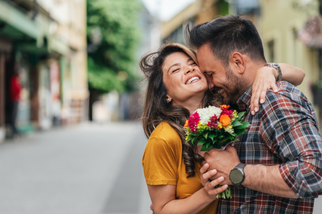 A smiling couple embracing while holding a bouquet of flowers, as if one partner has just given flowers to the other