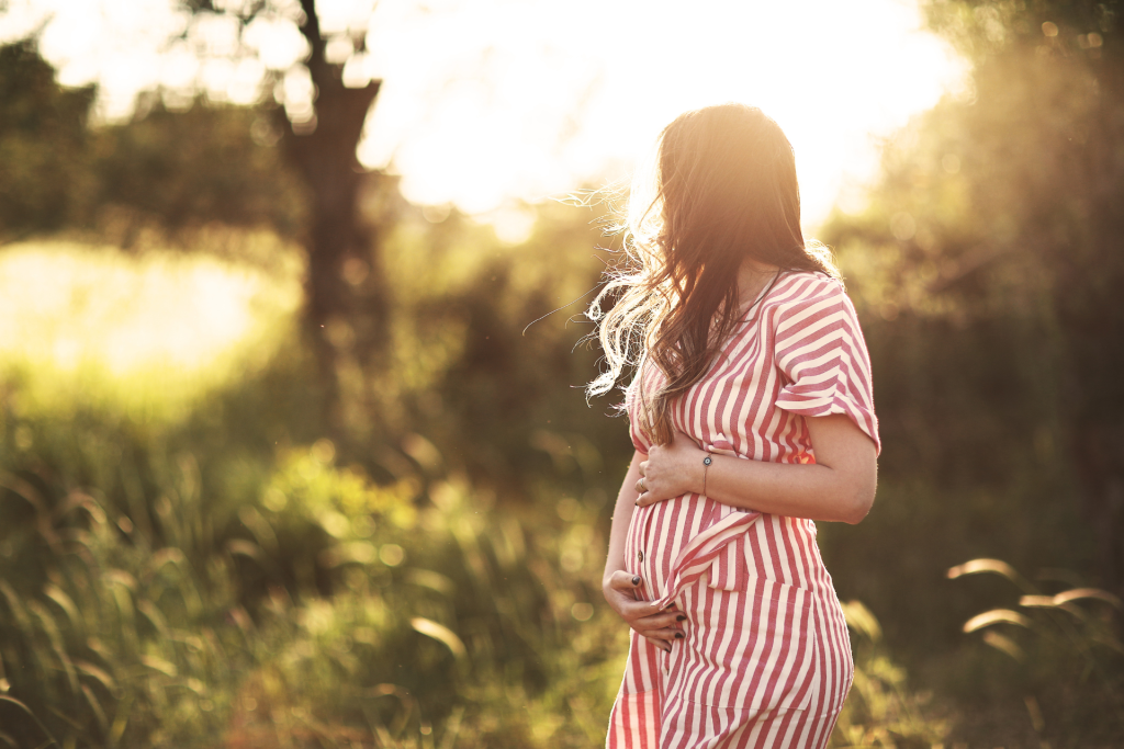 A pregnant woman walking outdoors at sunset, seen from the side and looking toward the horizon, with her face not visible