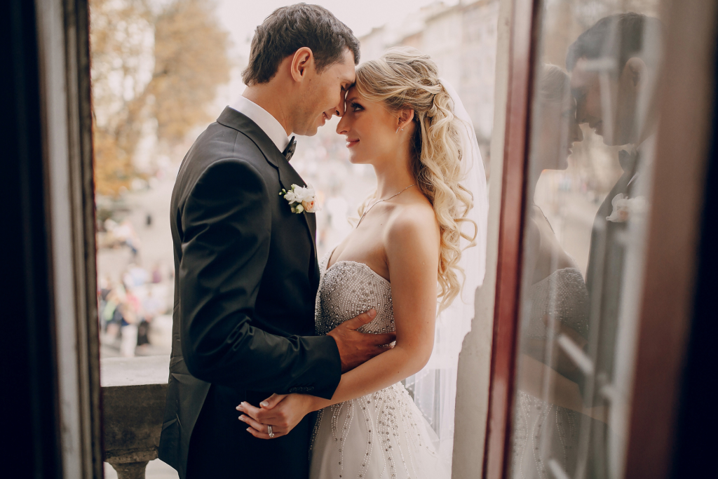 A bride and groom standing by a window on their wedding day, with the groom kissing the bride’s forehead as they both close their eyes