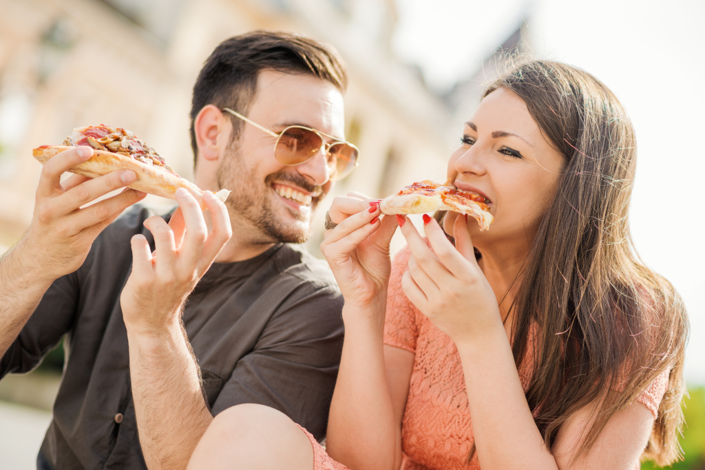A happy couple sitting together and eating pizza on a pleasant day, smiling and sharing a relaxed, affectionate moment