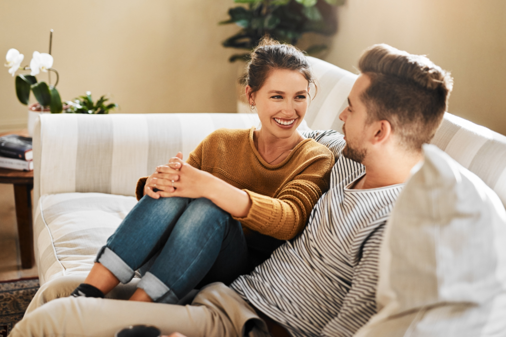 A couple sitting comfortably on a sofa with their feet up, smiling and looking into each other’s eyes while enjoying a calm, intimate conversation