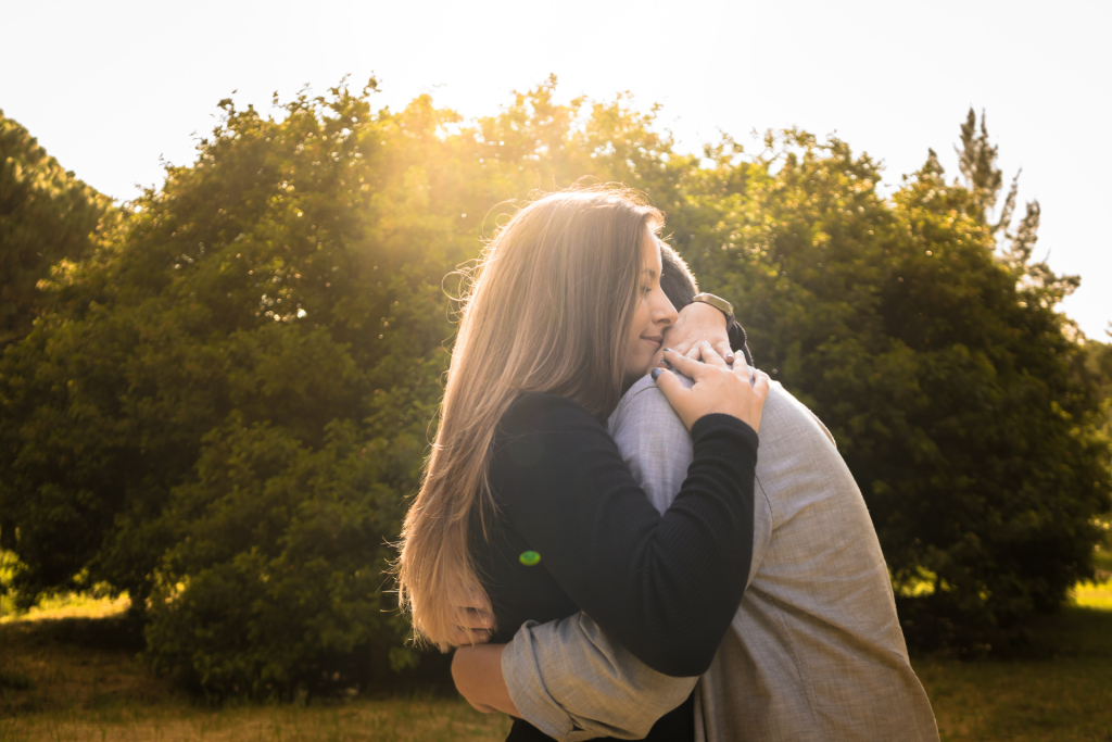 A couple embracing in a park on a sunny day, with trees and greenery visible in the background
