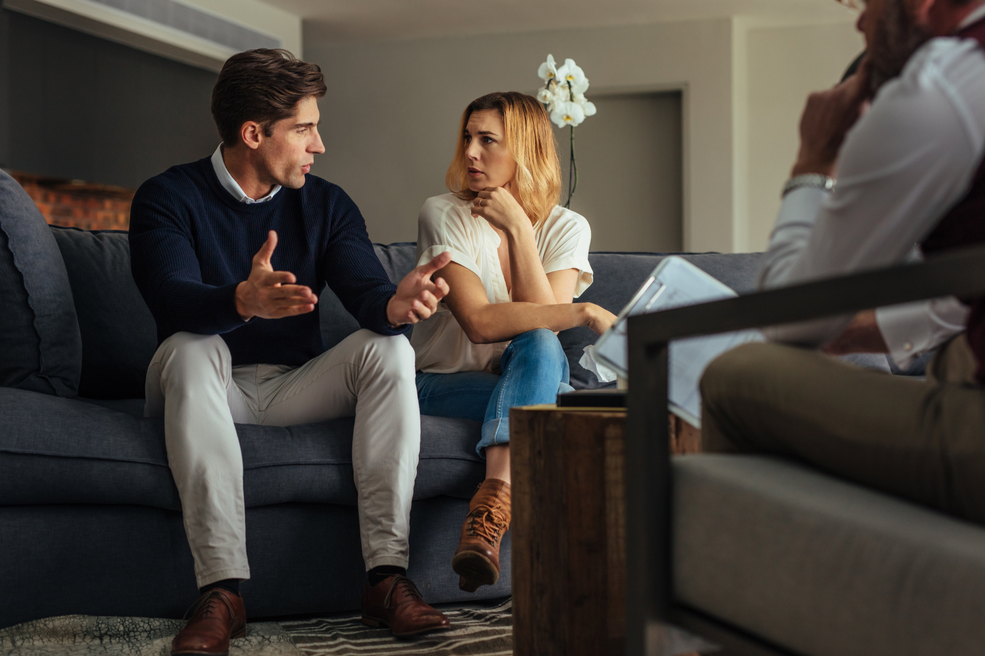 A couple sitting on a couch talking during a therapy session, with the therapist seen from behind as she listens to them