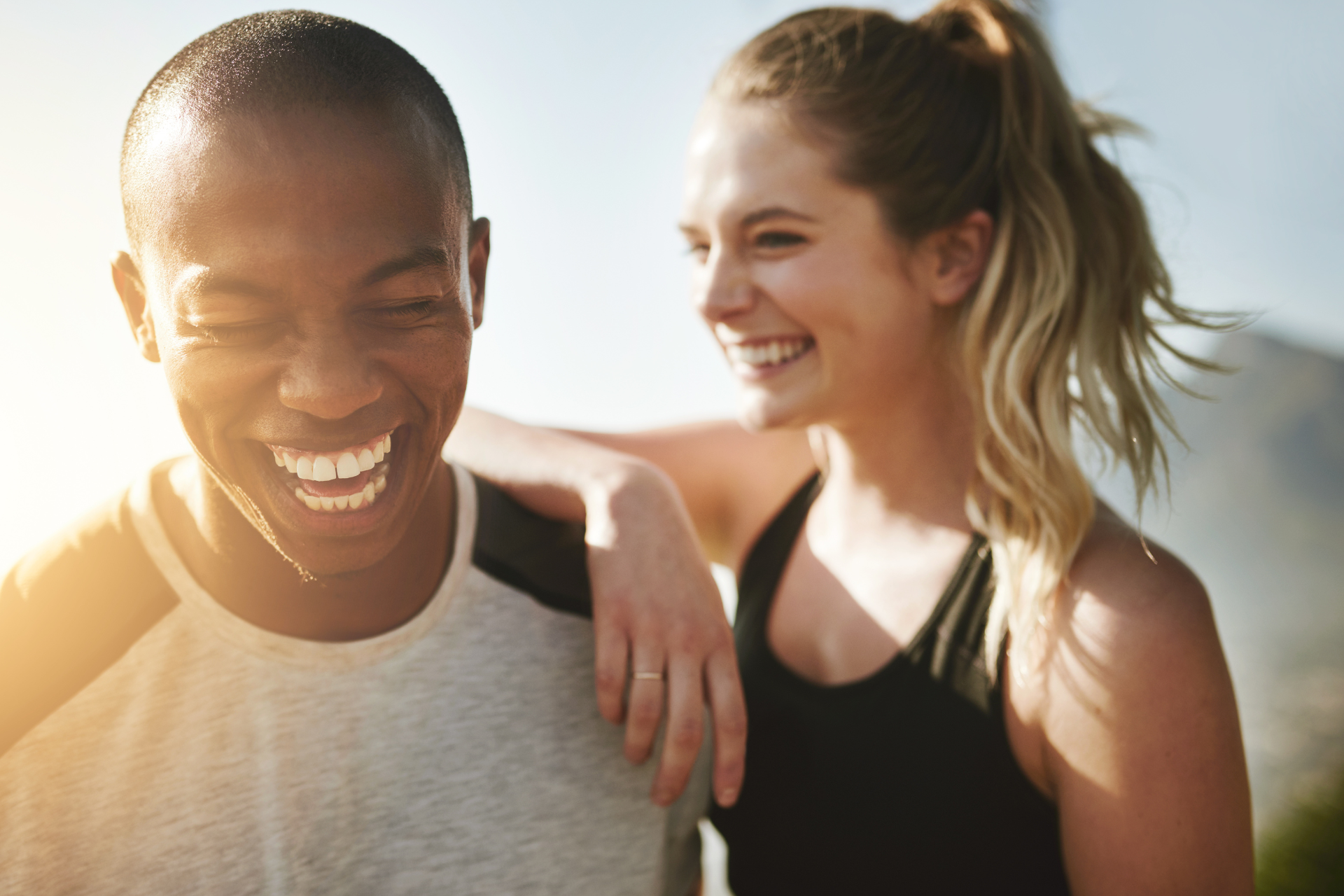A close-up of a man laughing with a woman smiling behind him, her hand resting on his shoulder, as they enjoy a sunny beach at sunset