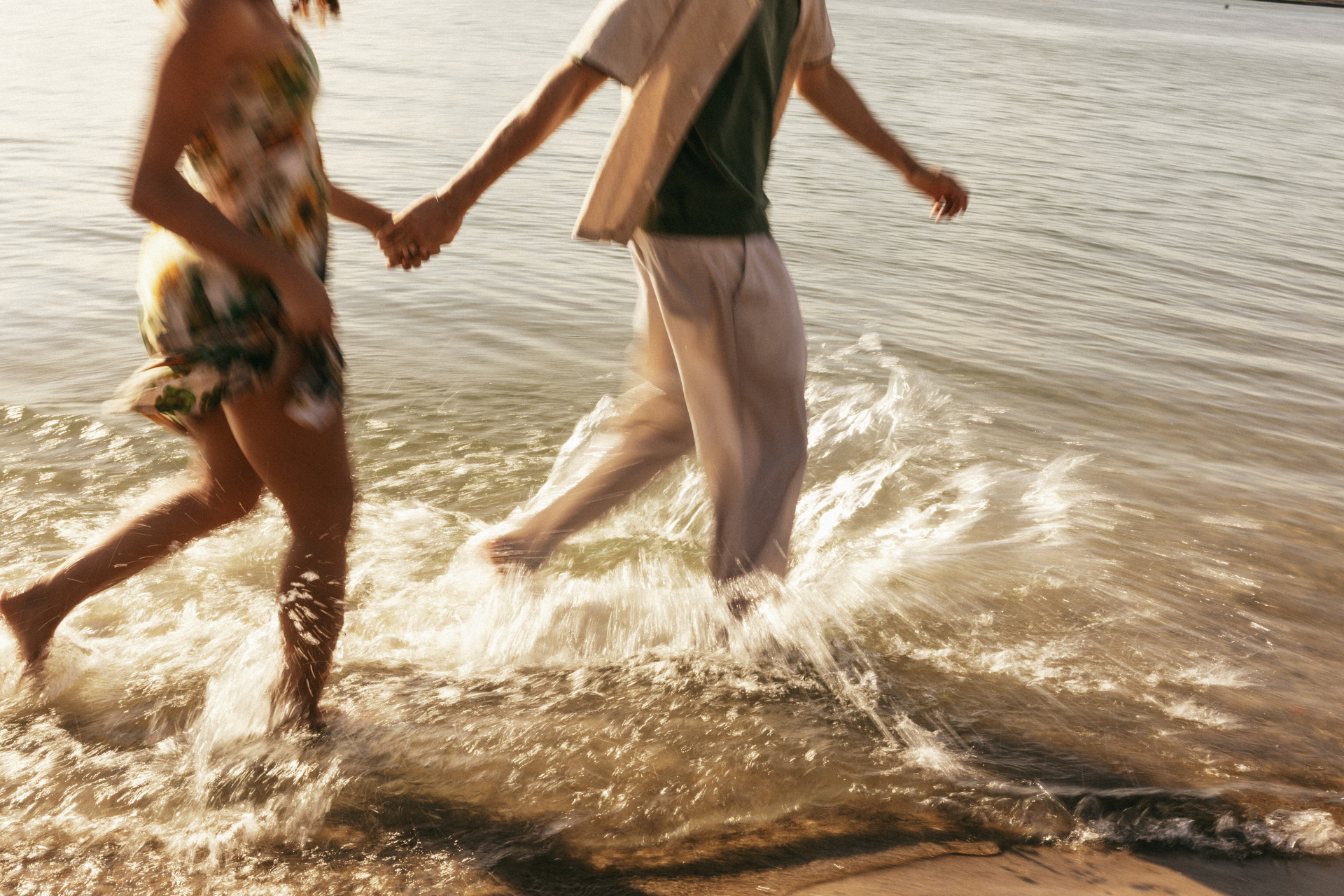 The legs of a couple running through shallow ocean water on the beach, holding hands and playing together at the shoreline