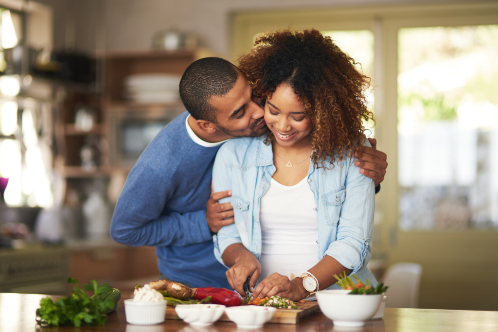 A woman chopping vegetables in the kitchen as her partner kisses her on the cheek, and she smiles during the affectionate moment