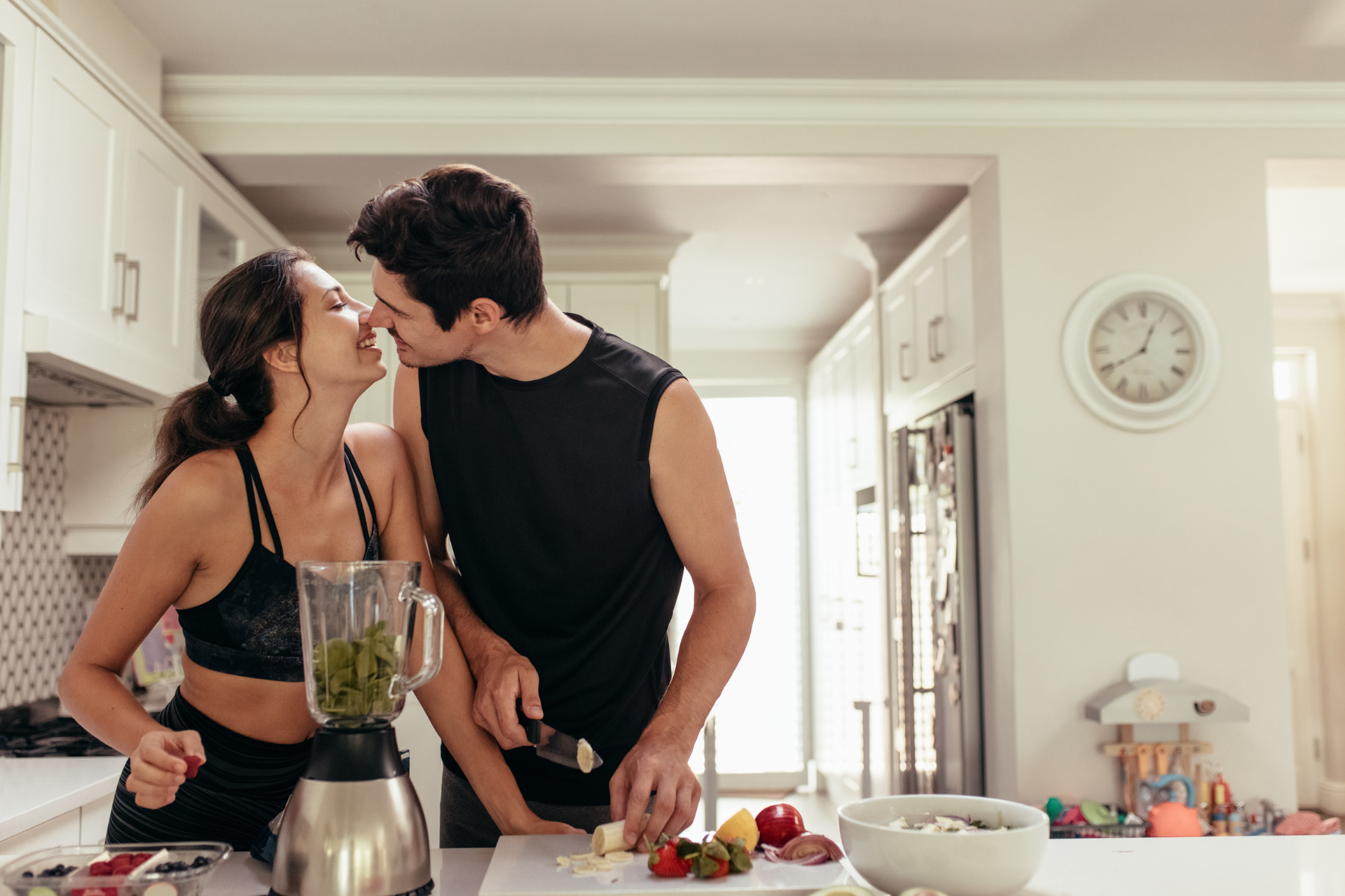 A couple kissing in the kitchen as the woman uses a blender and the man helps cut bananas and strawberries for a smoothie