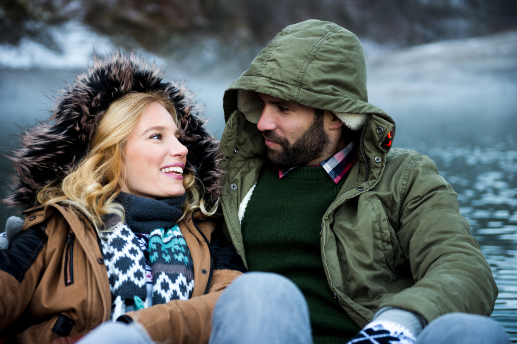 A couple gazing into each other’s eyes and smiling on a cold winter day, with snow and a lake visible in the background