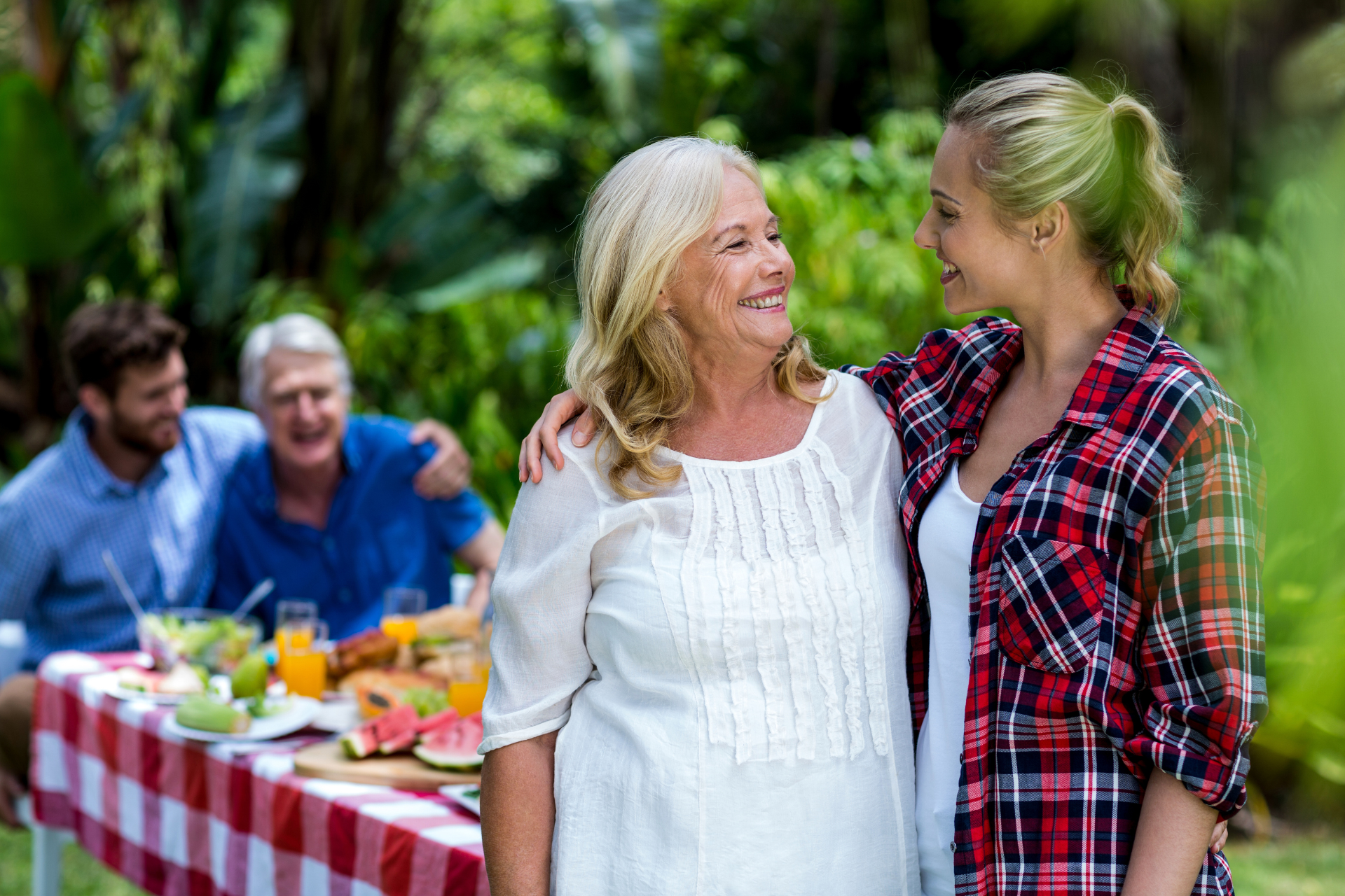 Family embrace during a gathering A close-up of a woman hugging another woman, appearing to be her mother-in-law, while in the background her partner stands with another man, possibly his father, during a family moment