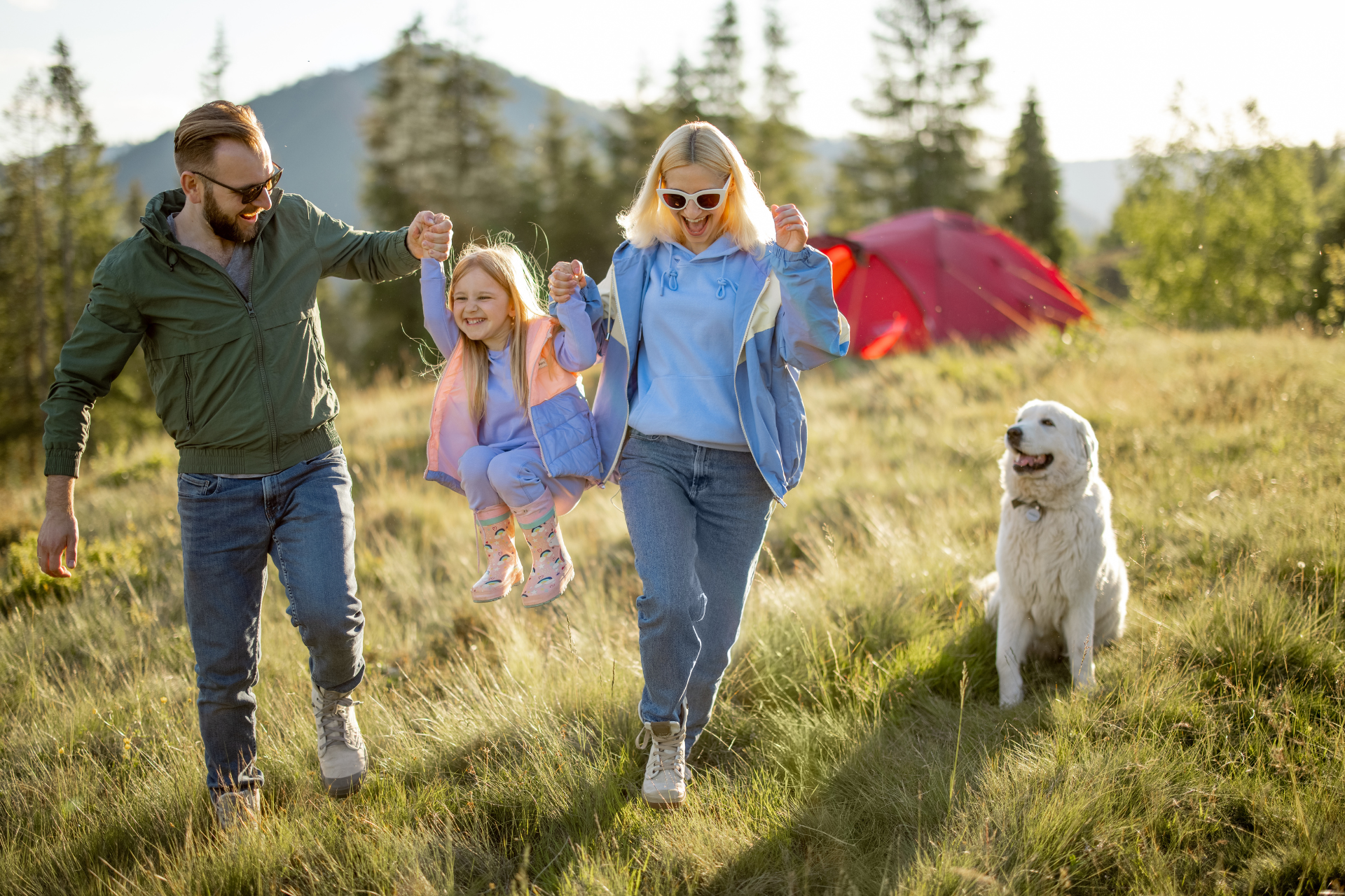 A mother and father lifting their daughter by the hands while laughing and playing outdoors, with their dog nearby enjoying the moment