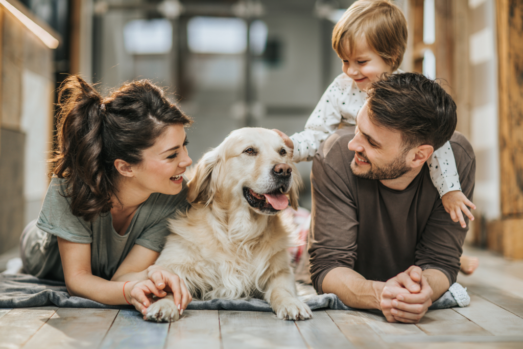 A family sitting on the floor with their dog in the center, as a child leans on the father and pets the dog while everyone looks at it affectionately