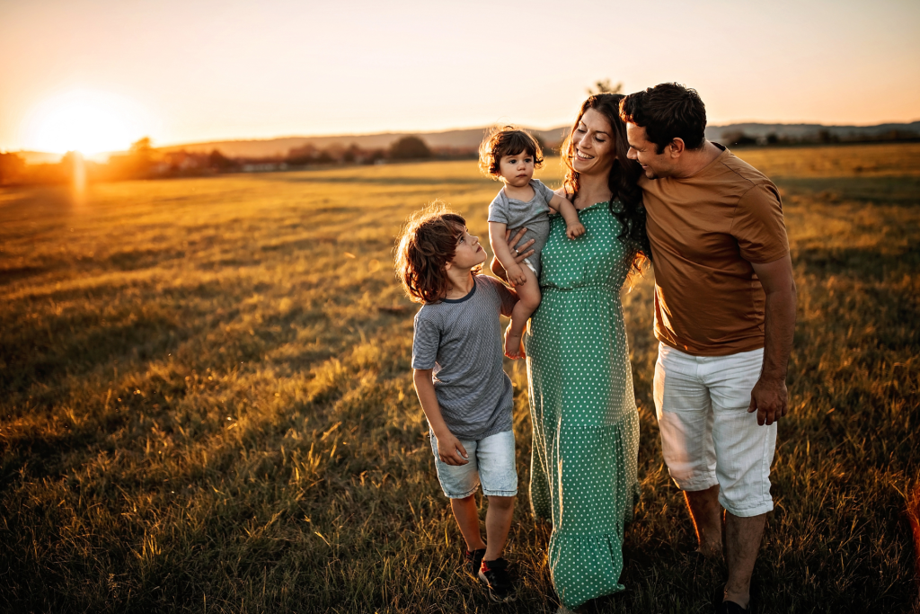A smiling family walking through a large field at sunset, with the mother carrying one child, another child beside her, and the father walking next to them in the warm evening light