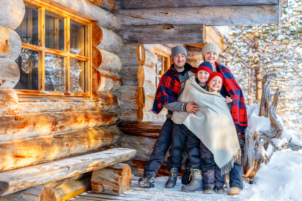 A mother, father, son, and daughter standing outside a cabin, wrapped together in a blanket, hugging and smiling at the camera