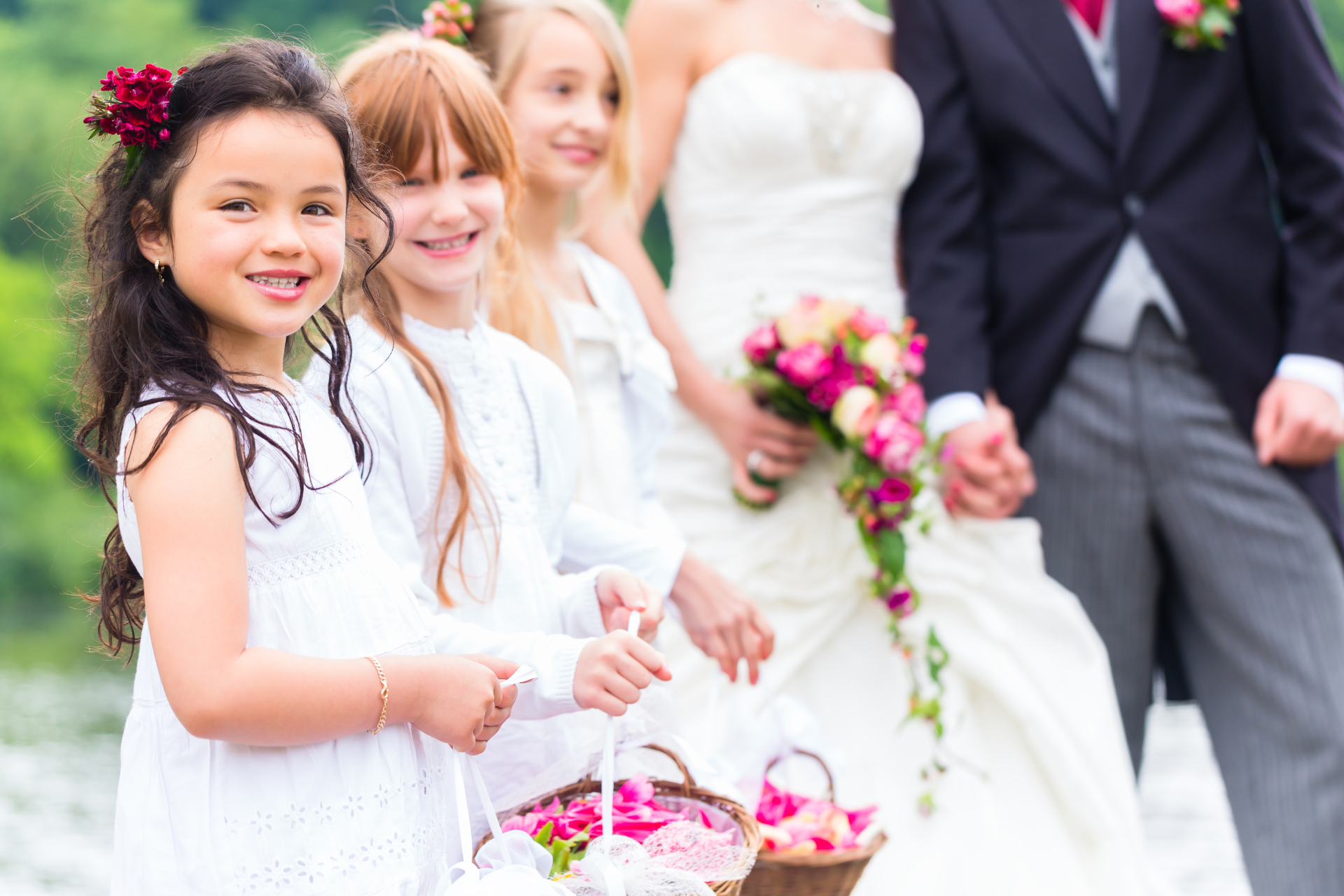 Several smiling flower children looking at the camera during a wedding, with the engaged couple blurred in the background behind them