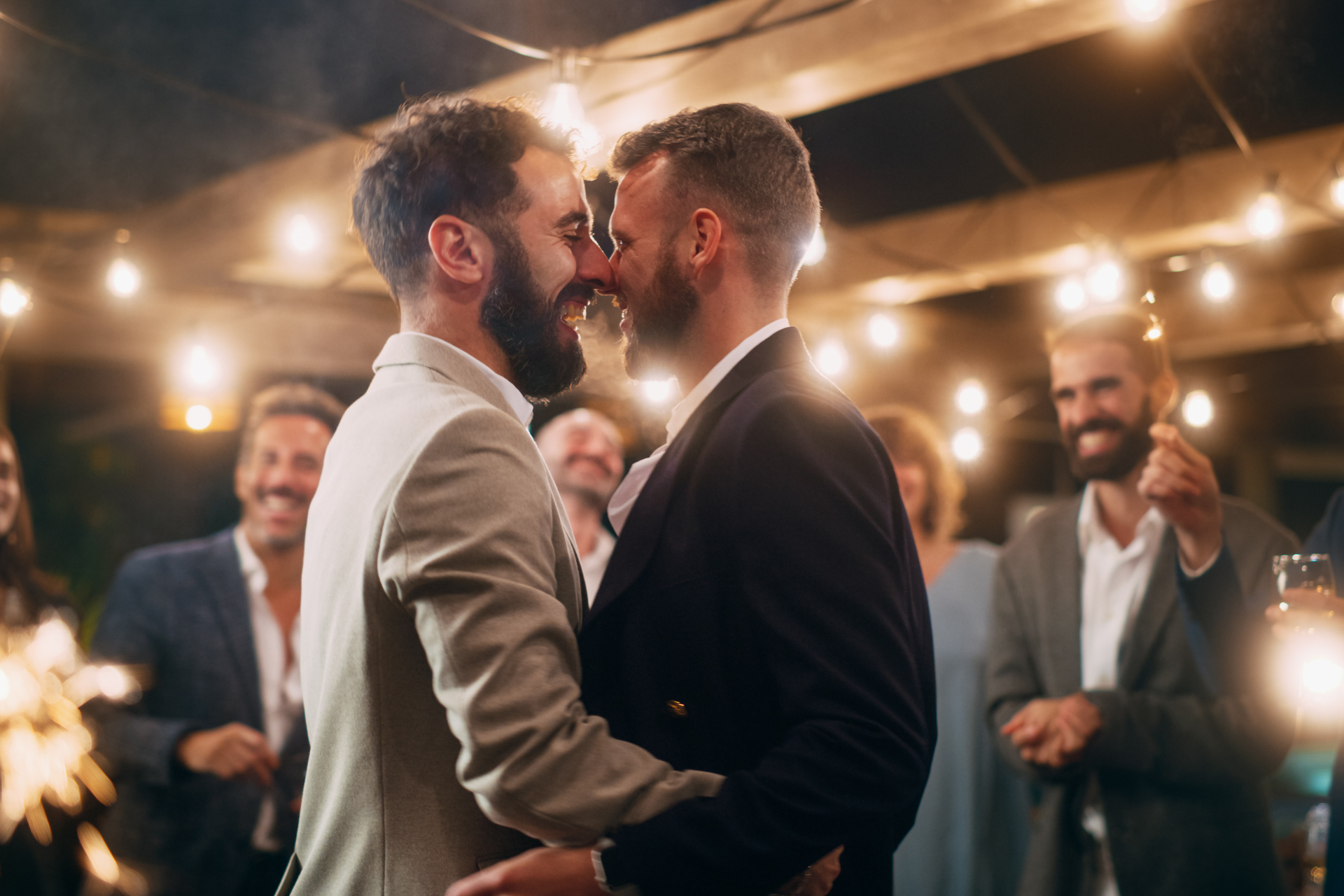 A joyful gay couple dancing together at their engagement celebration, with guests blurred in the background cheering and celebrating around them