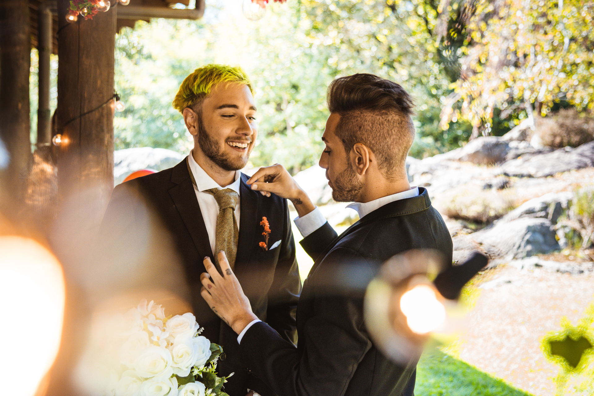 Two men at a sunny outdoor wedding helping adjust each other’s suits, sharing a quiet moment before the ceremony