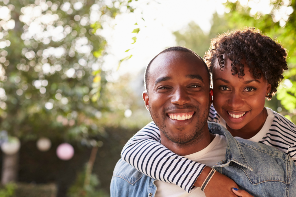 A smiling couple looking at the camera as the man lifts the woman, captured in a close-up outdoors with a softly blurred tree in the background