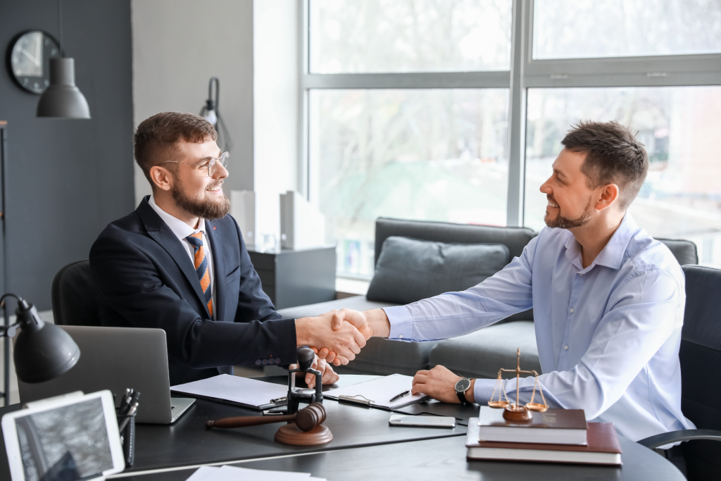 Two men smiling and shaking hands in an office setting, appearing to conclude a professional meeting between attorney and client